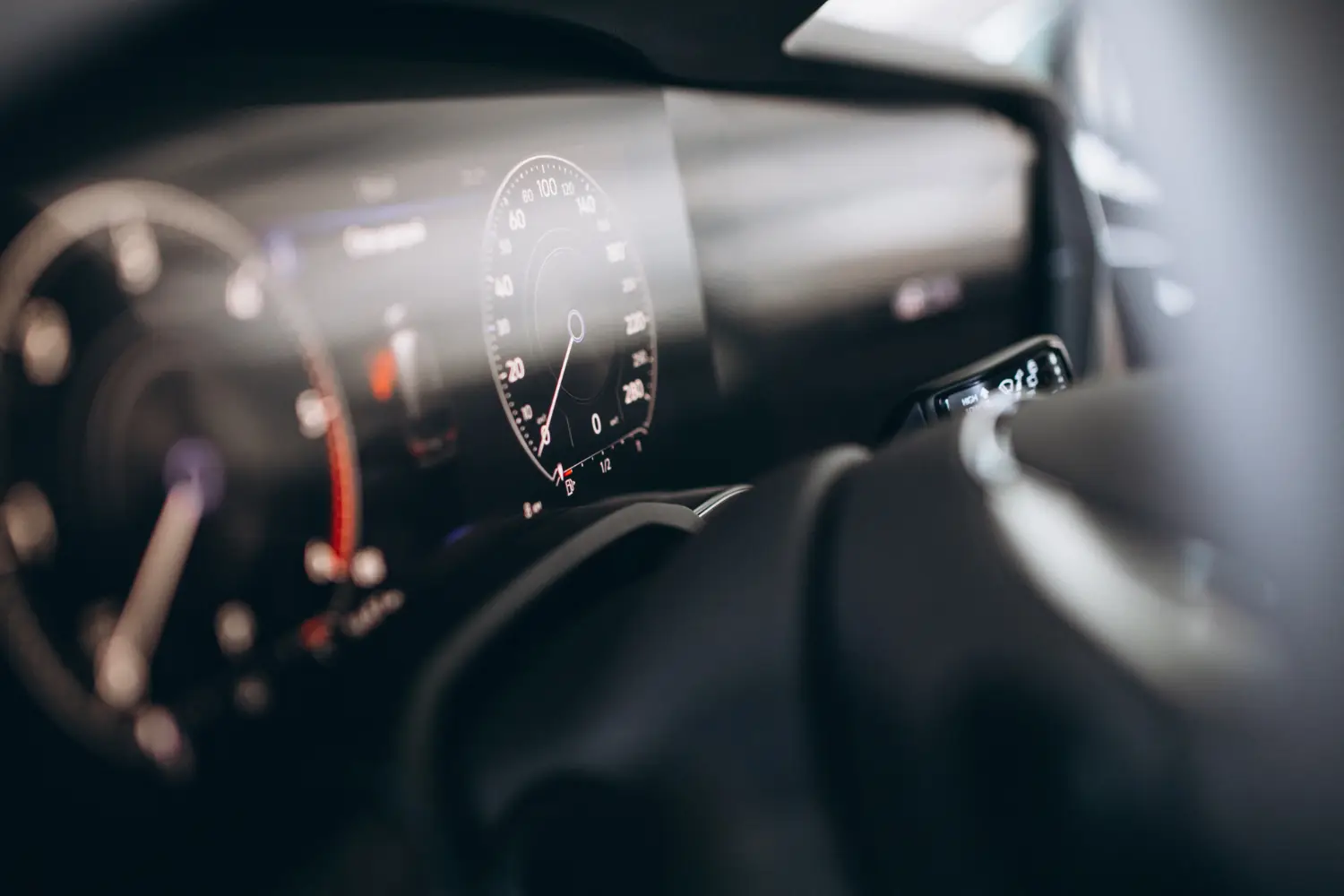 Eine detaillierte Nahaufnahme des modernen, dunklen digitalen Cockpit-Displays eines Fahrzeugs. Der zentrale kreisförmige Geschwindigkeitsmesser ist scharf fokussiert und zeigt eine große 'O' mit einem blauen Zeiger und Nummern von 20 bis 260. Andere Dashboard-Informationen und das Lenkrad sind weich unscharf im Vordergrund.